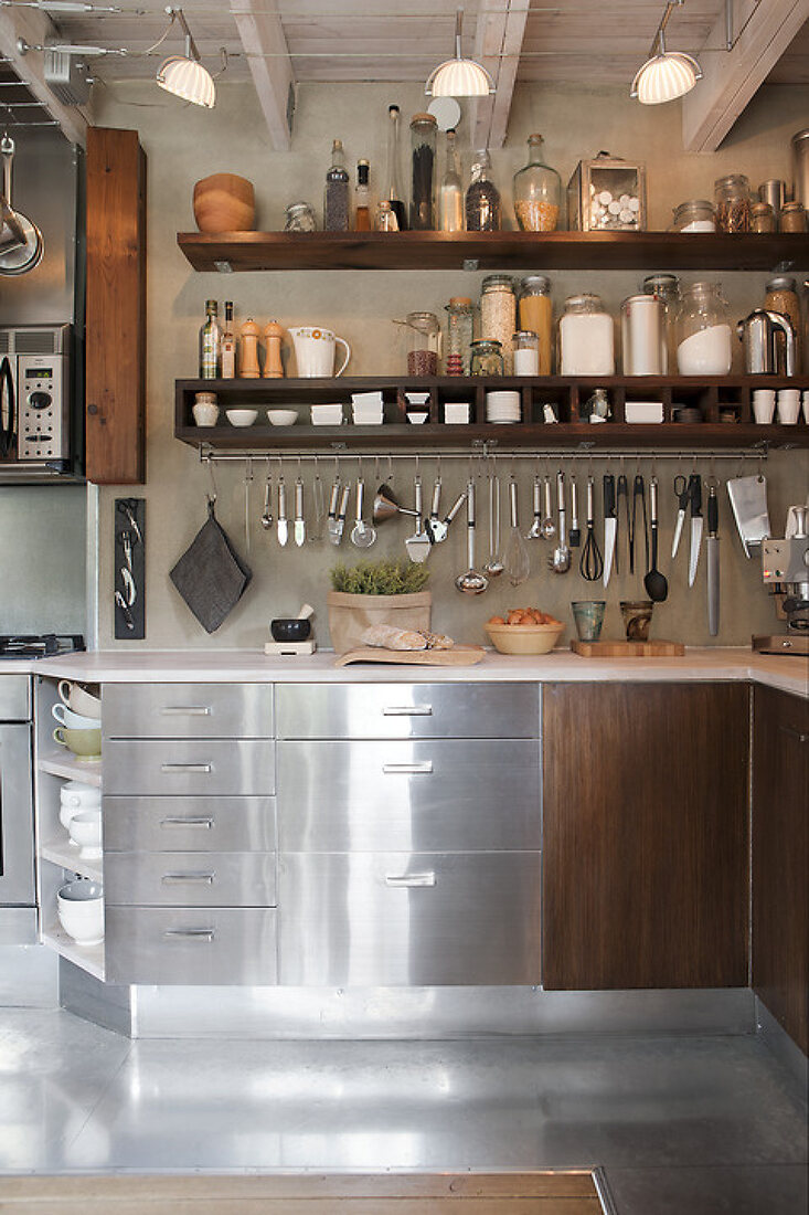 Kitchen utensils hanging from hooks on a long rail beneath open timber shelving above a metal worktop