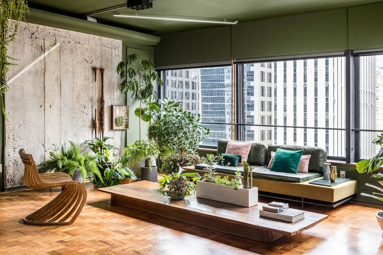 Long low timber coffee table with planters, surrounded by abundant indoor plants beside large living-room windows