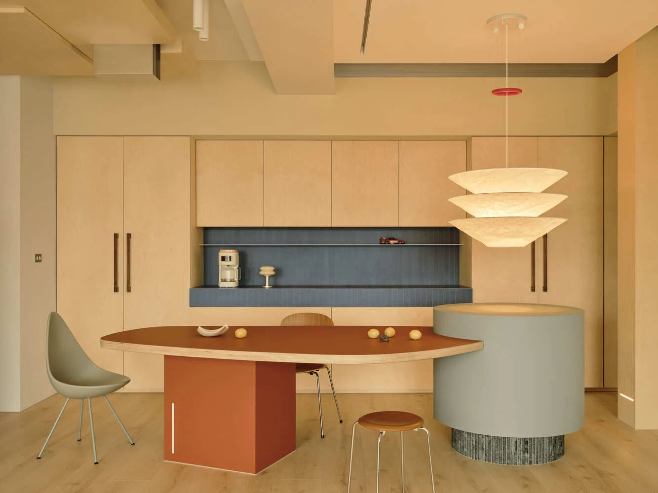 Pale timber kitchen cabinetry, blue recessed worktop and sculptural dining table beneath a coordinated timber ceiling